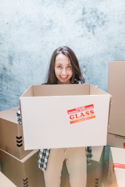 Close-up of a person wearing a dark green shirt and dark trousers holding a large cardboard moving box with both hands. The box features a red and white sticker reading 'CAUTION THIS SIDE UP' with an