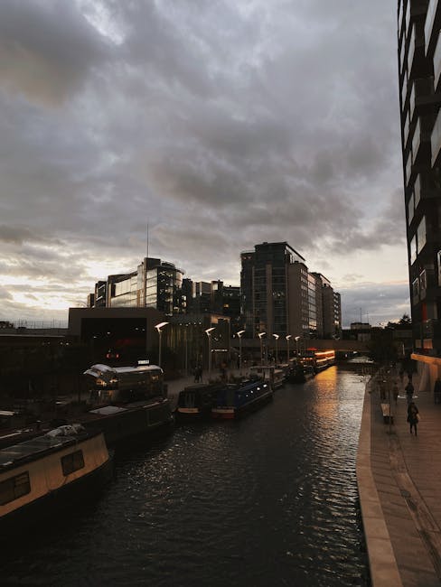 A view of Regent's Canal in Haggerston during late afternoon or early evening, with dark, overcast clouds covering the sky and a faint hint of sunlight on the horizon. Modern multi-storey buildings line the canal with reflective glass windows, and several parked boats are moored along the water's edge. On the right side of the image, a paved walkway curves alongside the canal, with pedestrians walking along it. The scene is illuminated by soft ambient light, and the urban environment suggests a busy area suitable for efficient home relocation and furniture transport. This setting is consistent with the surroundings of a house removals service, such as those provided by Man with Van Haggerston, supporting the logistics of packing, loading, and transit along key routes in the Haggerston area.