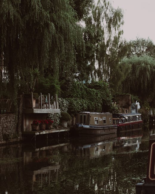 A canal scene along Regent's Canal in Haggerston during daylight, featuring a blue and white houseboat moored on the right side of the waterway, with visible safety ropes and life rings on its roof. On the left, a traditional brick building with large windows and a modern, multi-story structure with extensive glass panels overlook the canal. A large tree with lush green foliage partially obscures one side of the scene, casting shade onto the walkway and the houseboat. The water in the canal is calm, reflecting the sky and surrounding buildings, which are under a partly cloudy sky with patches of blue. The image captures an environment suitable for home relocation or furniture transport activities, with the presence of a boat potentially used for moving supplies or equipment, and the urban setting indicating proximity to residential or commercial properties. Occasionally, there are people visible in the background near the water's edge, engaging in activities related to planning or preparation for moving.