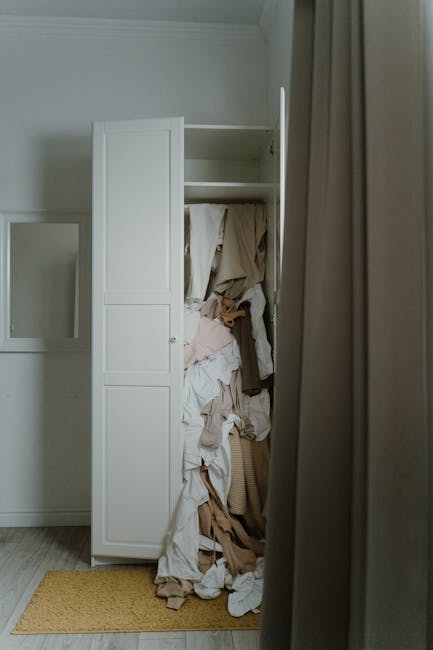 A tall, white, partially open wardrobe placed against a white wall in a room with wooden flooring. The wardrobe's door is ajar, revealing a cluttered interior filled with crumpled and hanging clothing items in various neutral tones such as beige, white, and light pink. Next to the wardrobe, a beige curtain partially covers the view, with a small yellow mat on the floor beneath. The scene appears to be part of a house being prepared for moving or packing, consistent with a home relocation process supported by Man with Van Haggerston, who offers professional removals and furniture transport services.