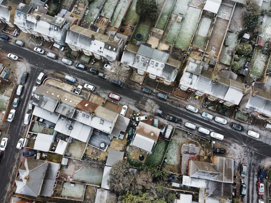 An aerial view of a residential neighbourhood showing a narrow street lined with parked cars and closely spaced terraced houses with pitched roofs, some covered with roofing materials and roof vents. The houses have small backyards with fences, gardens, and sheds. In the foreground, a home undergoing a furniture transport or packing and moving process is visible, with a partially opened vehicle nearby. The driveway and pavement areas contain cardboard boxes, wrapping materials, and furniture being loaded or unloaded, with visible blankets and straps used for securing items during transportation. The surroundings are consistent with an urban setting, and the lighting suggests daytime with overcast weather. This scene reflects the typical environment for house removals and home relocation services, such as those offered by Man with Van Haggerston, highlighting logistical details like furniture loading, packing materials, and street-level movement relevant to house removals on Graham Road Estate.