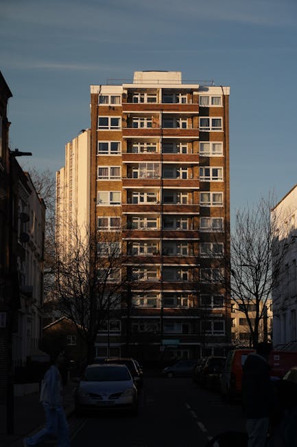 A multi-storey residential building with a brick and concrete exterior, featuring numerous balconies with glass railings and large windows, standing on a street lined with leafless trees and parked vehicles including a white car in the foreground. The scene is captured during daylight with clear skies overhead. In the foreground, two individuals are walking along the pavement, and the base of the building suggests an urban environment suitable for house removals or furniture transport services. The photograph emphasizes the high-rise nature of the property and the surrounding street scene, aligning with home relocation and moving logistics, as seen on the service page for house removals by Man with Van Haggerston.