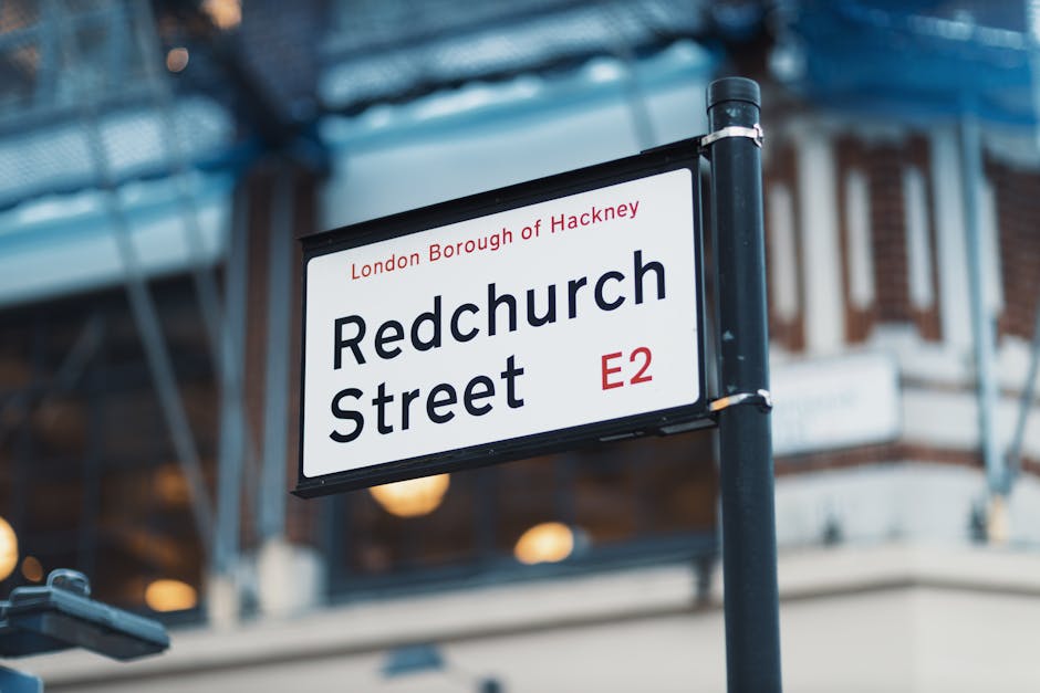 A street sign mounted on a black pole displaying 'Redchurch Street' in bold black letters, with the smaller text 'London Borough of Hackney' and the postcode 'E2' in red. The sign is situated outdoors on Redchurch Street in Hackney, with a building featuring brick and white facade details visible in the background. The image is well-lit, capturing the sign and surrounding environment during daytime. This scene relates to the local area where house removals and move planning, including obtaining parking or moving permits from Hackney Council, may be necessary, a service offered by Man with Van Haggerston during home relocations.