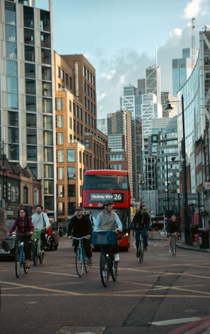A busy urban street scene in Haggerston with several pedestrians and cyclists navigating the roadway, including a group of four cyclists in the foreground, one of whom is carrying a large blue package on his bike. Behind them, there is a red double-decker bus displaying route information for Hackney Wick, number 26, moving through the street surrounded by tall modern office and residential buildings made of glass, steel, and brick. Vehicles are parked along the pavement, and some pedestrians are walking on the sidewalk. The sky above features a mix of clouds and sunlight, illuminating the scene evenly. This image captures an active moment in city life that relates to the logistics of house removals and transport services, with potential reference to local permits or moving logistics in Haggerston, managed by Man with Van Haggerston.