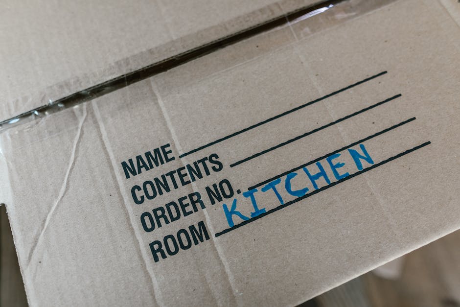 Two women engaged in a home relocation process indoors, surrounded by stacked cardboard moving boxes of various sizes, some sealed with red packing tape. One woman, with shoulder-length brown hair, is