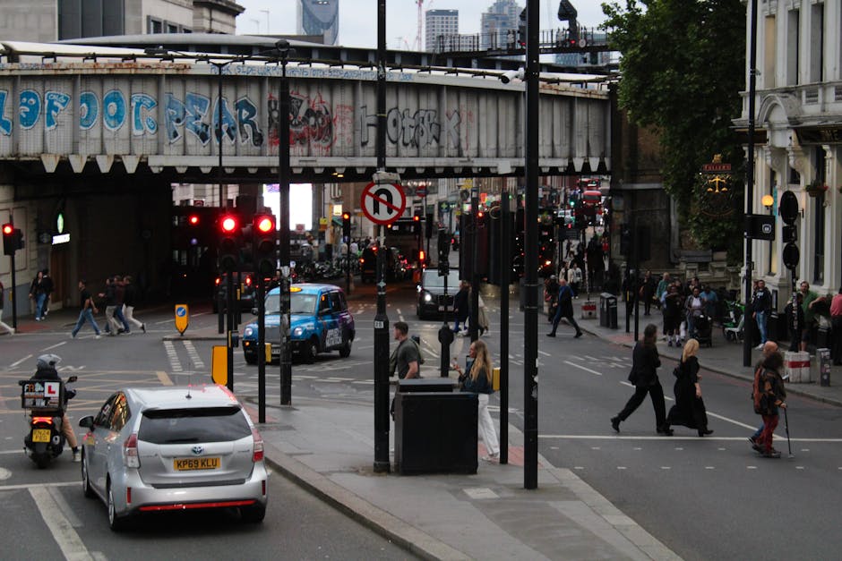 A busy urban intersection in Haggerston during daytime, featuring pedestrians crossing the street and waiting on the pavements, with vehicles including a silver Toyota Prius and a blue van moving through the intersection. Overhead, a concrete railway bridge with graffiti spans across the roads, supported by vertical columns, and traffic lights, street signs, and lamp posts are visible at the corners. The scene includes buildings with shops and residential properties on both sides, and some shops have illuminated signs. The area appears to be part of a home relocation or furniture transport process where the street environment is active with city traffic and foot traffic, with [COMPANY_NAME] operating in the background as part of the moving services for residents or businesses on Kingsland Road near Haggerston Station.