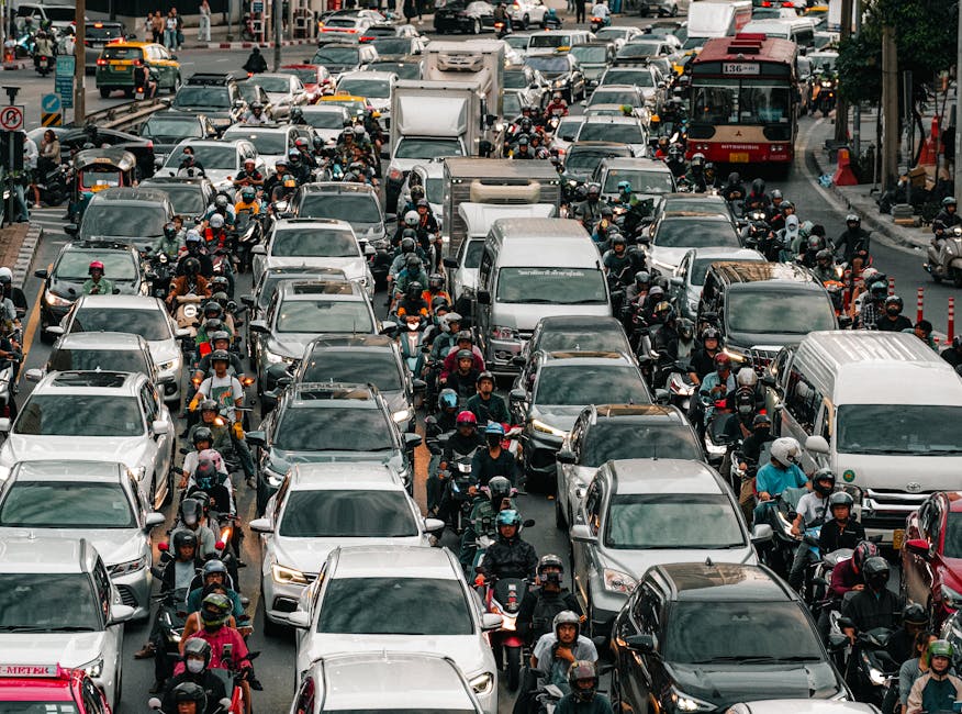 A street scene showing heavy traffic congestion with numerous cars, including taxis and private vehicles, occupying all lanes of the road in front of a row of multi-storey residential buildings. Some cars are closely packed and appear stationary, reflecting typical urban traffic during peak hours. In the foreground, a person wearing a black helmet and a dark jacket is walking beside a silver SUV, which is in the process of being loaded or unloaded as part of a home relocation or moving process. The scene is outdoors during daylight with clear weather, and the background reveals a mix of urban infrastructure, traffic signs, and greenery. This image is relevant to house removals and furniture transport services provided by Man with Van Haggerston, illustrating the busy environment encountered during local moving logistics.