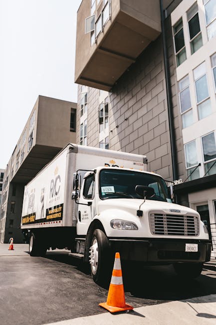 A white box truck with the logo 'RDC' on the side, parked outside a modern multi-story building with beige and white facade, during daylight hours. The truck is positioned on a paved driveway, with orange traffic cones placed in front of it to mark the loading area. The truck's side door is closed, and there are no visible personnel. The building features large windows and protruding sections, indicating contemporary architecture. This scene captures the logistics involved in home relocation, with the truck likely used for furniture transport and packing material movement as part of a house removals service, supporting moving and packing processes typical of professional removals companies like Man with Van Haggerston.