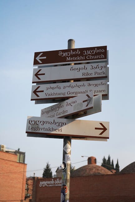 A tall metal street sign pole displaying multiple directional signs in both Georgian and English, indicating destinations such as Metekhi Church, Rike Park, Vakhtang Gorgasali Square, the Botanical Garden, and Leghvtakhevi. The signs are made of metal with a weathered appearance, with some pointing left and others pointing right, mounted at different heights on the pole. The background features a clear sky, the tops of trees, and residential rooftops, suggesting an urban setting. This type of signage may be encountered in areas offering guidance for visitors exploring Haggerston or similar neighborhoods, supporting navigation during home relocation or moving processes.