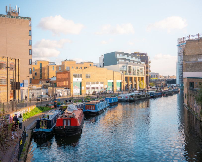 A daytime scene along a narrow canal in Haggerston, showing multiple narrowboats docked side by side with their brightly painted exteriors reflected in the water. On the left, a group of people is standing on a paved walkway near the edge of the canal, some engaging in conversation. Behind them, a metal fence separates the walkway from a building with a chain-link gate. The buildings lining the canal include modern residential apartments with large windows and balconies, as well as older brick industrial-style structures. The sky above is partly cloudy with sun illuminating the scene, emphasizing the vibrant colours of the boats and the surrounding architecture. This environment is typical for urban house removals, where loading and unloading of furniture and boxes may occur near the canal – a service provided by Man with Van Haggerston for home relocations or furniture transport needs.
