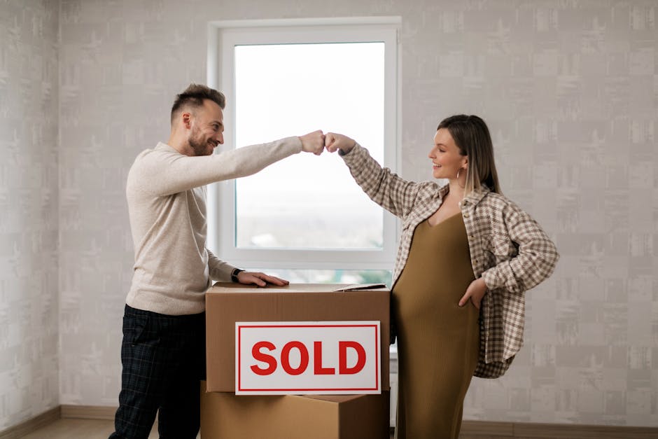 A man and a pregnant woman indoors, standing behind a cardboard box marked with a red 'SOLD' sign, during a home relocation process. The man, dressed in a light sweater and checkered trousers, is smiling as he extends his right fist forward towards the woman. The woman, wearing a plaid shirt over a brown dress, is also smiling and is reaching out her right fist to meet his for a fist bump. The scene takes place in a room with a large window allowing natural light to fill the space, revealing plain wallpapered walls and a minimal interior. The box is situated on a wooden floor, and the window behind them suggests a daytime move involving packing and moving tasks, consistent with furniture transport and home relocation services provided by Man with Van Haggerston, specialists in removals and moving logistics.