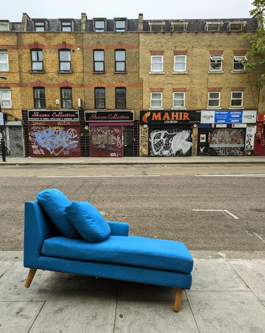 A bright blue chaise lounge with two matching cushions is positioned on the sidewalk outside a commercial building in Haggerston. The furniture, covered in fabric, stands on wooden legs and faces the street, suggesting it has been removed from a nearby property as part of a house relocation or furniture transport process. Behind the chaise, the building features a row of shopfronts with closed metal roller shutters painted with graffiti art and tags. The shop signs include one for 'Sharma Collection' and another for 'Mahir London' with additional signage for professional services. The street is empty of traffic, and the overall scene indicates an ongoing or completed move, with the furniture temporarily placed outside. The image is well-lit with natural daylight, emphasizing the urban environment typical of London neighborhoods like Haggerston, and illustrating the logistics involved in packing and moving household items for short-term storage or relocation handled by [COMPANY_NAME].
