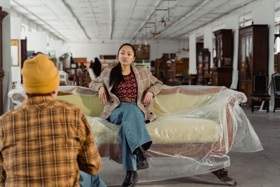 A woman with long dark hair, dressed in a checked blazer, red patterned shirt, and blue jeans, is seated on a piece of furniture covered with protective bubble wrap in an indoor furniture warehouse. She is positioned in the center of the image, with her legs crossed and her left arm resting on the furniture, displaying a neutral expression. In front of her, a person wearing a yellow beanie and a plaid shirt is kneeling, facing her, likely engaged in a conversation related to the home relocation process. The warehouse environment features a concrete ceiling with exposed pipes and beams, and an array of antique wooden cabinets, dressers, and chairs arranged in the background. Some of these furniture items are partially wrapped for protection, suggesting they are being prepared for transportation or storage. The scene depicts the process of packing, handling, or planning furniture transport during a move, with the workspace lit by natural and artificial light, reflecting cautious and organized moving procedures, as managed by Man with Van Haggerston.