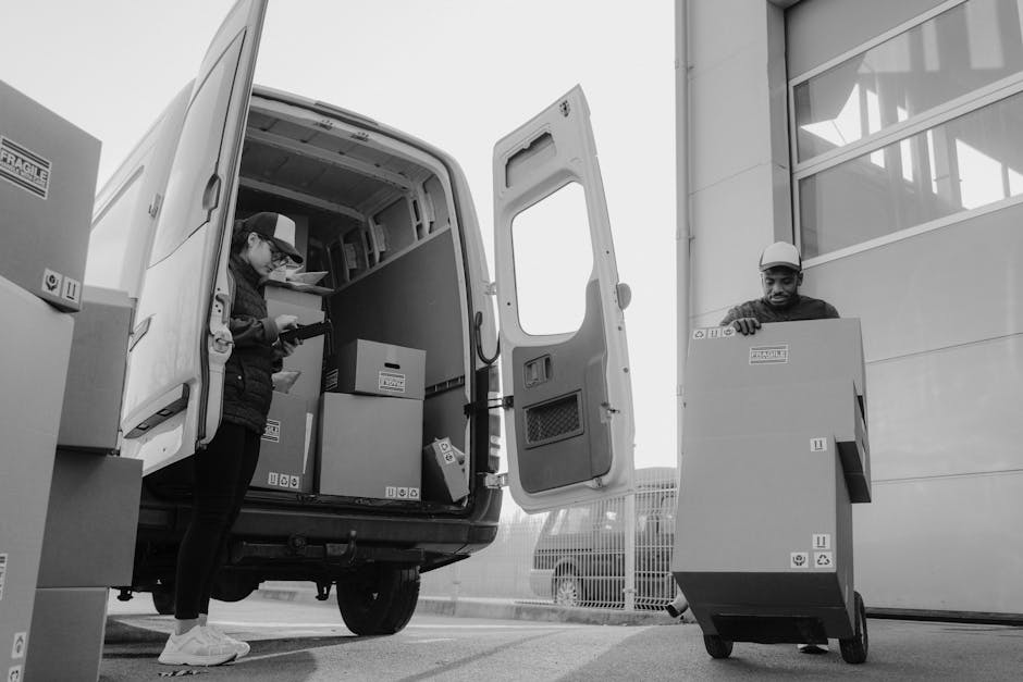 Inside a white moving van with open rear doors, three movers are preparing for a home relocation in Haggerston. The van's interior contains several cardboard boxes, some wrapped in plastic or fabric, 